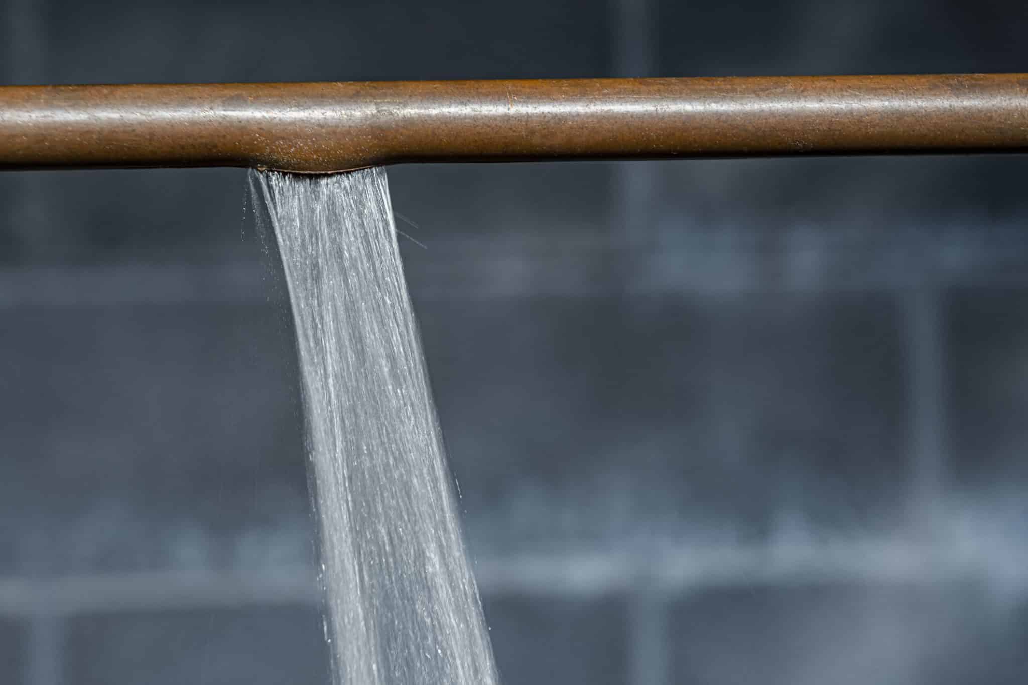 A horizontal copper pipe, spraying water against a blurry brick background.