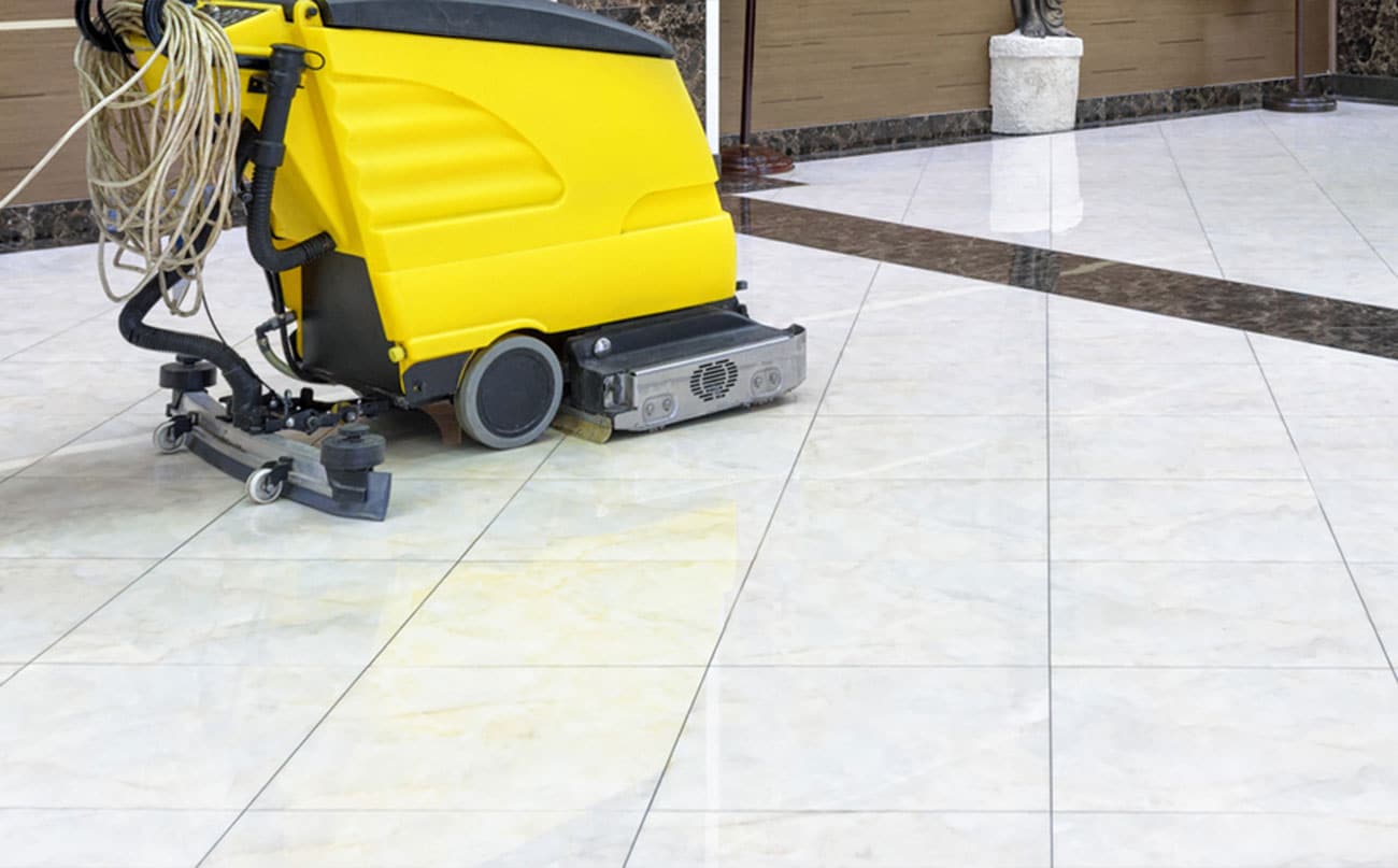 A person in blue pushes a floor cleaner down a wide hall in an academic building